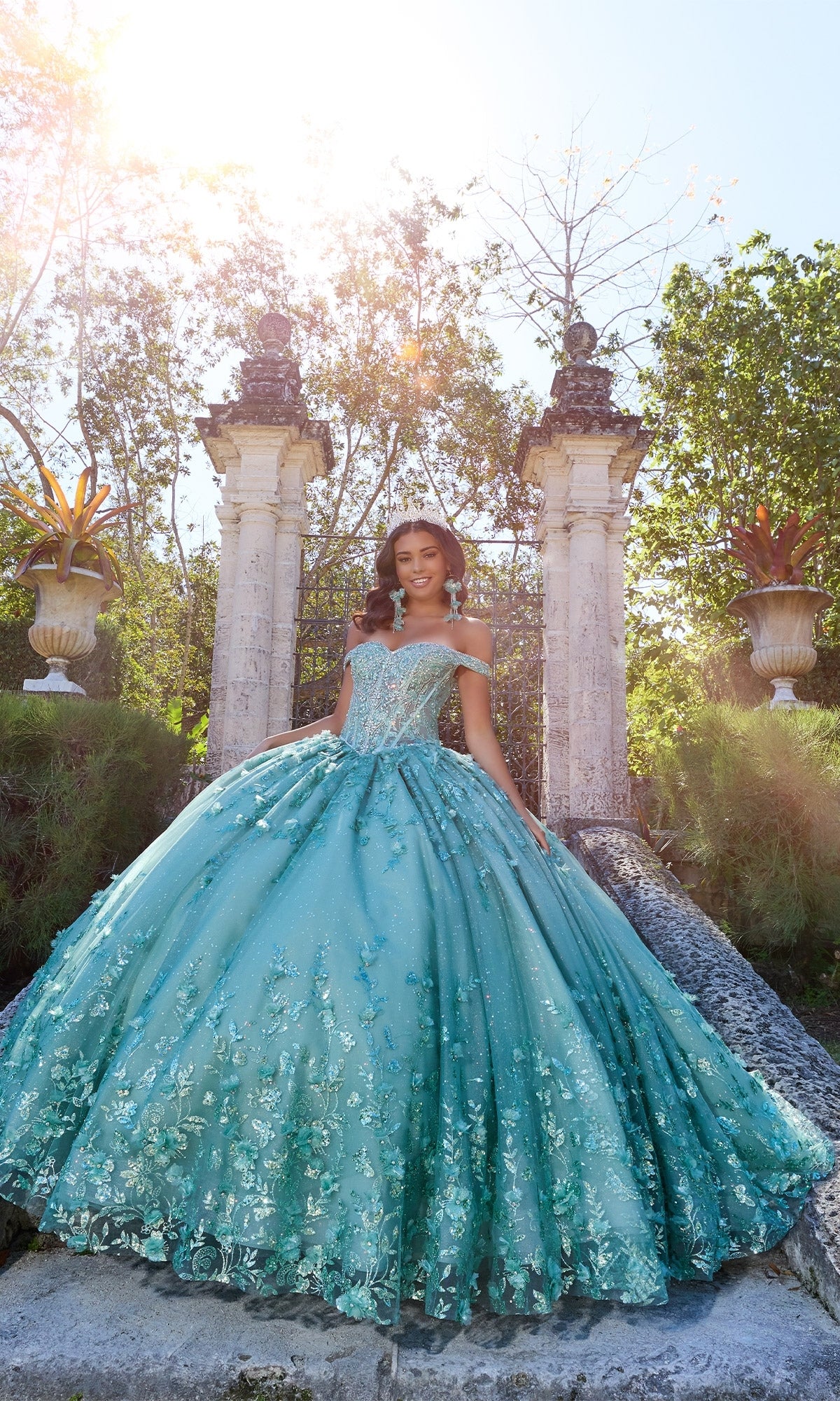 A woman in the Princesa PR30131 off-shoulder quinceanera dress with 3D floral appliques stands outdoors before stone columns and lush greenery, sunlight shining from above.